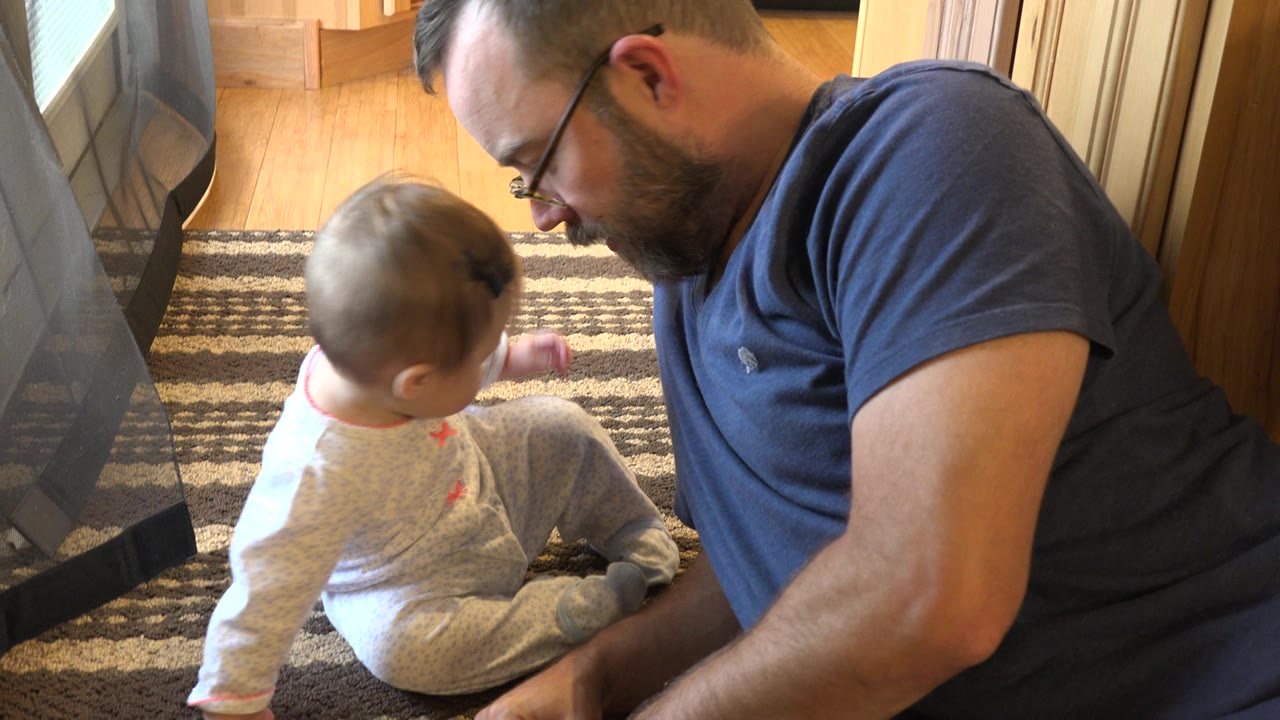 Father lying beside baby seated on a striped rug near a glass door.