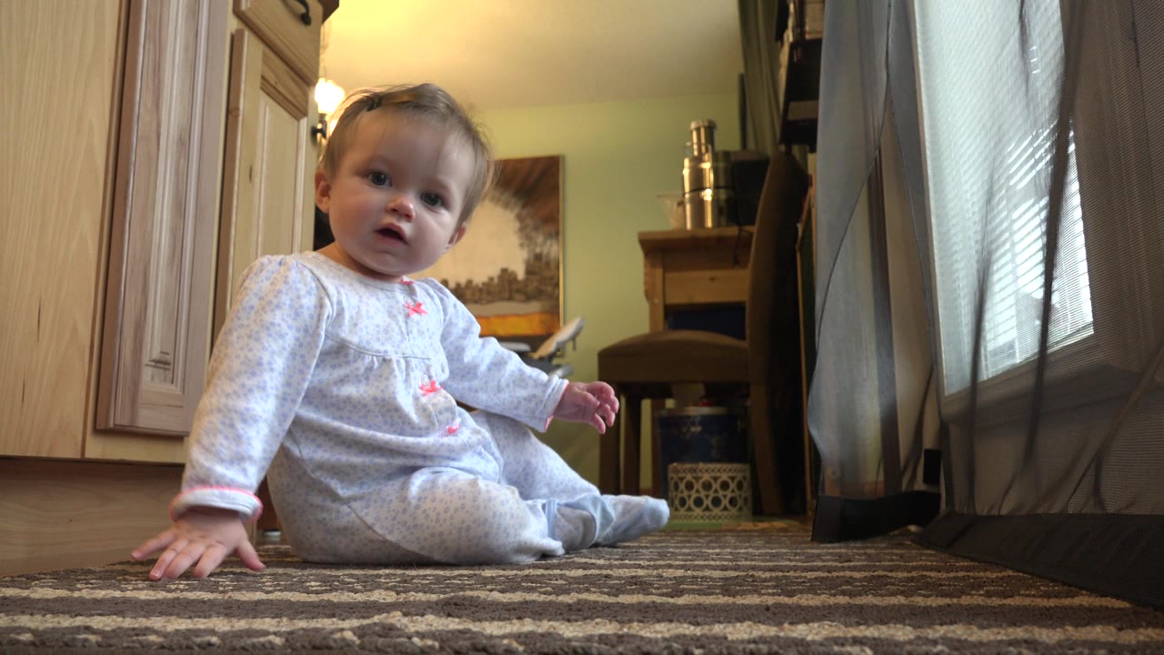 At 0:01, the baby leans to one side with a hand on the rug while the wood cabinetry and sheer curtain remain visible behind.