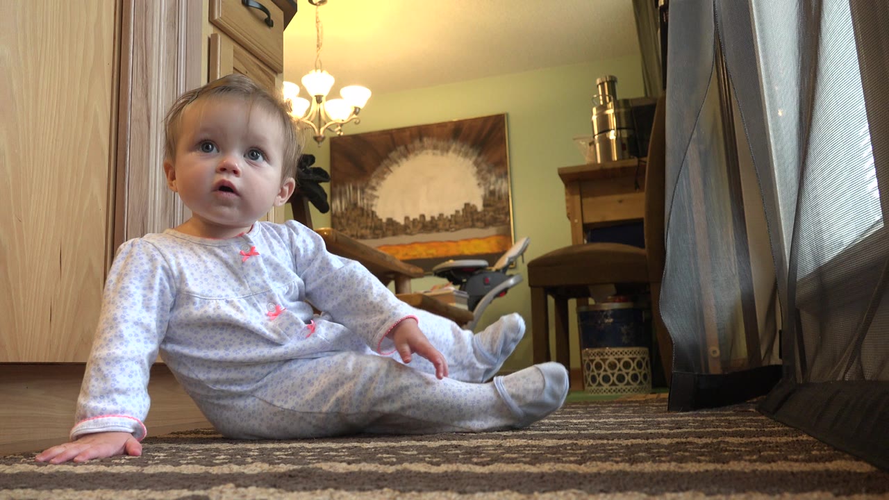 Baby girl in light pajamas sits on a striped carpet in a Westminster, Colorado home, with wood cabinetry, curtains, warm ceiling light, and wall artwork behind her.