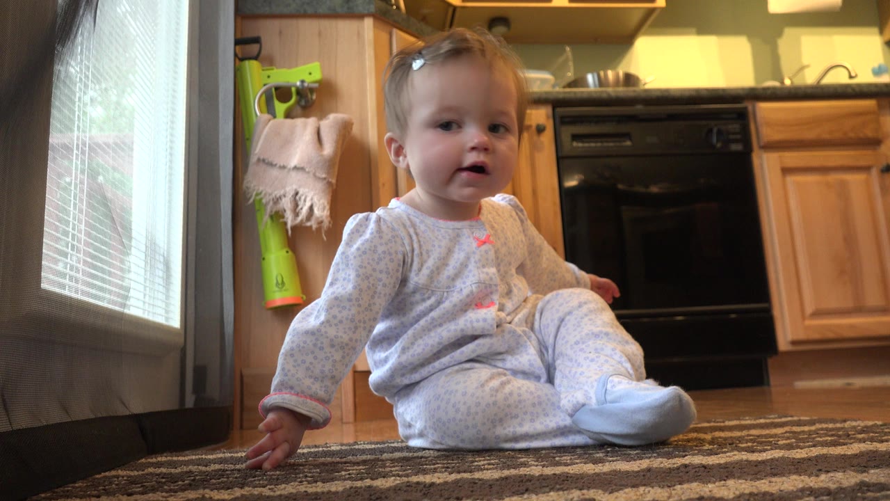 A toddler in light pajamas sits facing the camera on a patterned rug inside a Westminster, Colorado kitchen, with wood cabinets and a black oven behind her.
