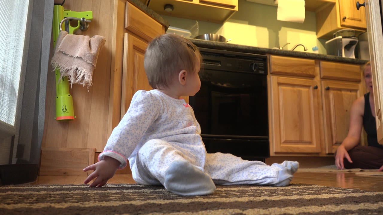 At 0:00 in Westminster, Colorado, a toddler in light pajamas sits on a patterned rug near wooden kitchen cabinets while a mother is partly visible at the right edge.
