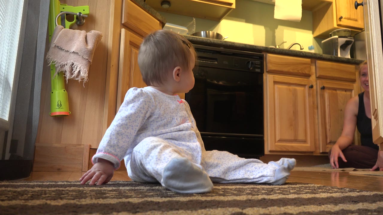 At 0:02 in Westminster, Colorado, the toddler sits slightly turned toward the mother, with warm cabinets and a dark kitchen appliance behind them.