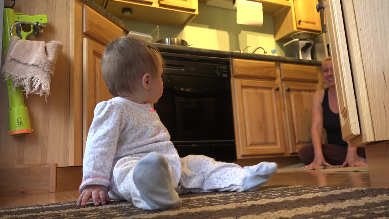 Baby girl seated on a kitchen rug with an adult partly visible in the background.