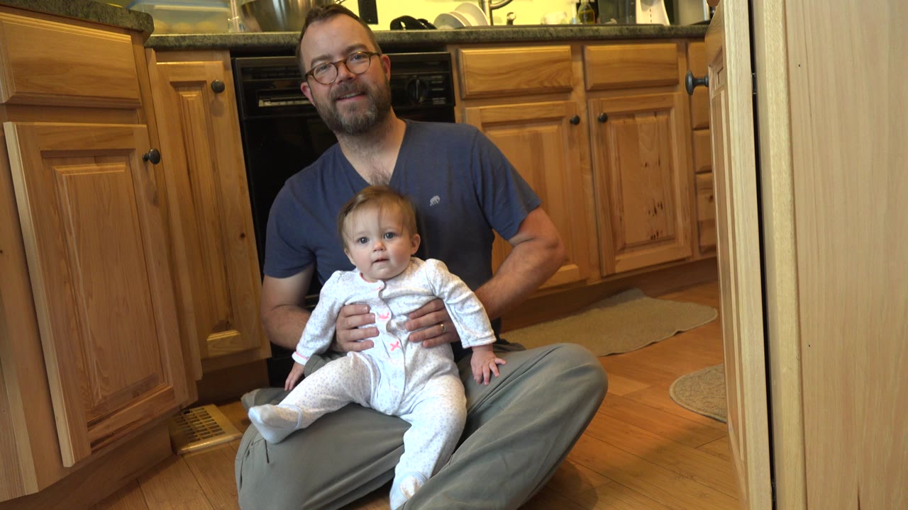 In a Westminster, Colorado kitchen, a bearded man in glasses sits on the wood floor holding a baby girl in a white sleeper in front of wood cabinets.