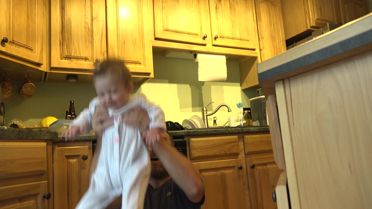 In Westminster, Colorado, the toddler remains lifted near the center of the kitchen frame while the father's face is mostly below the child's body.