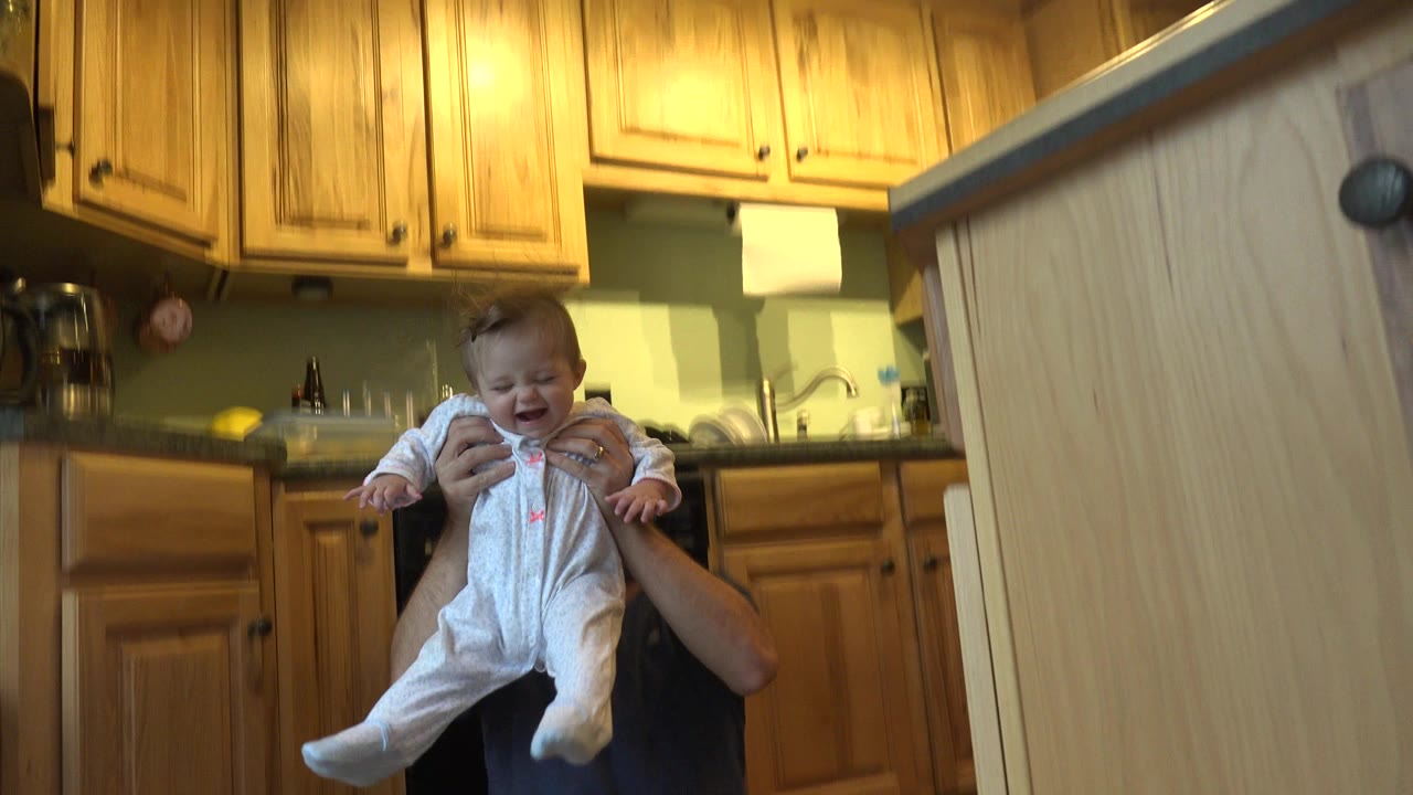 In a Westminster, Colorado kitchen, a father partially hidden behind a foreground cabinet lifts a baby in a white sleeper beneath warm wood cabinets.