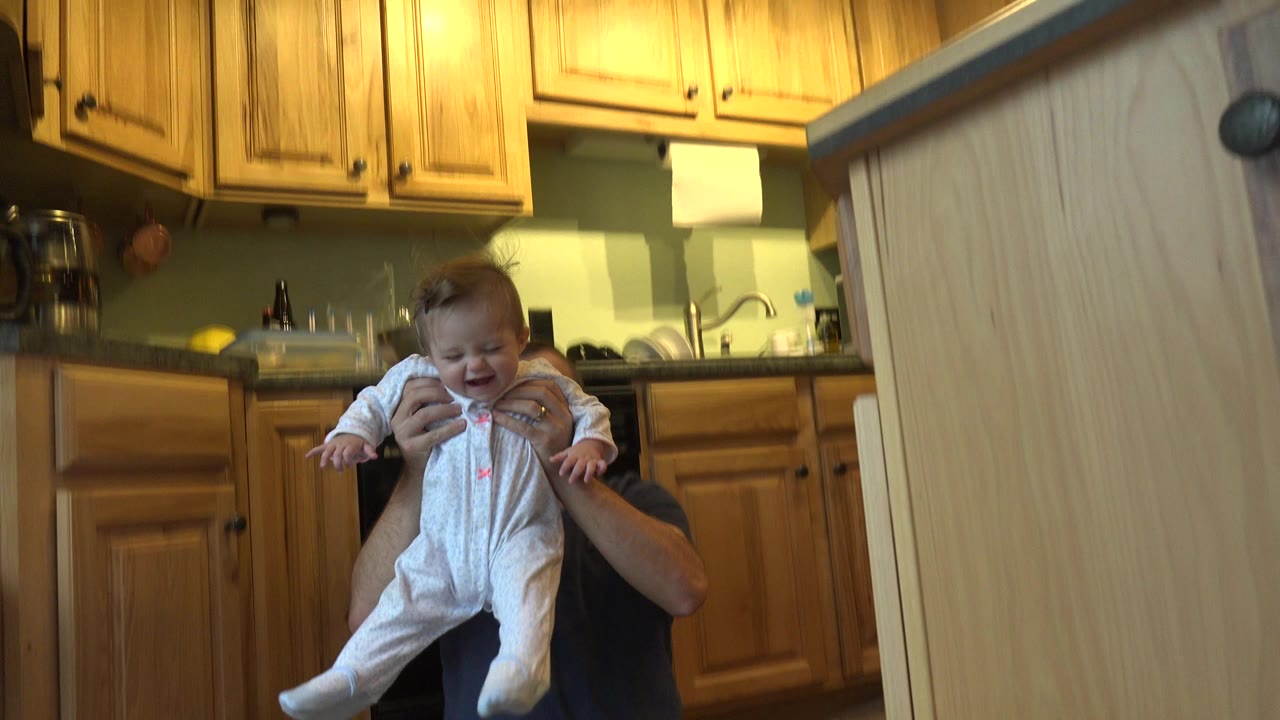 A baby in a white sleeper is held higher near the center of a Westminster, Colorado kitchen, with the father's head mostly obscured behind a cabinet edge.