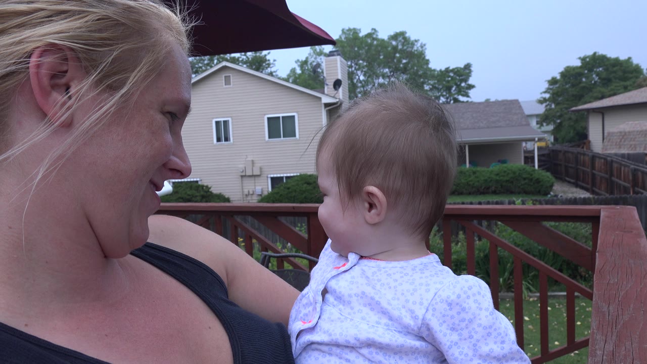 The mother smiles down at the baby girl as they face each other on the Westminster deck, with beige houses and green trees in the background.