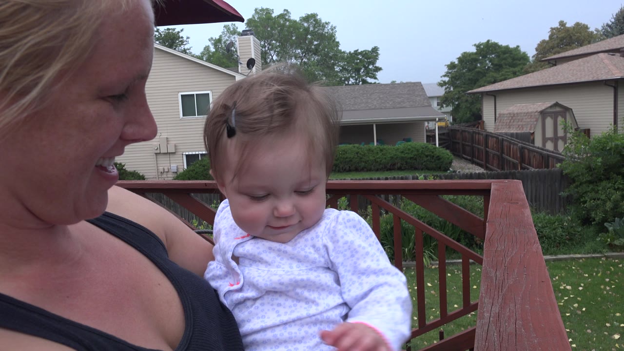 The baby looks downward in the mother's arms on the Westminster deck, with a red railing and beige houses in the background.