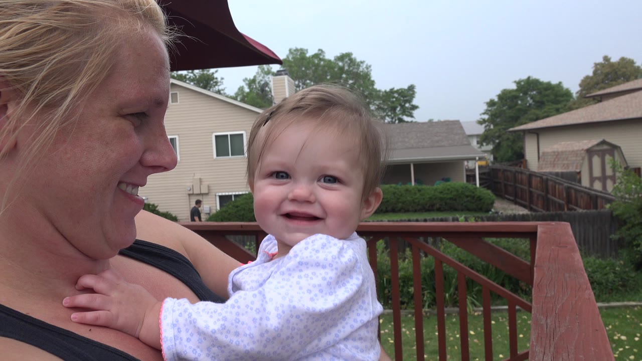 In Westminster, Colorado, a blonde woman holds a smiling baby beside a red wooden deck rail, with suburban homes and trees behind them.