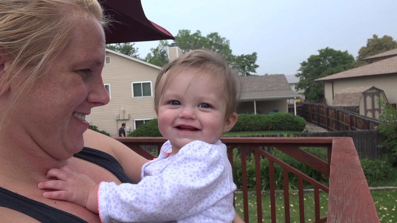 In Westminster, Colorado, the woman smiles at the baby as the child faces the camera, with the red rail, fenced yard, and nearby houses in the background.