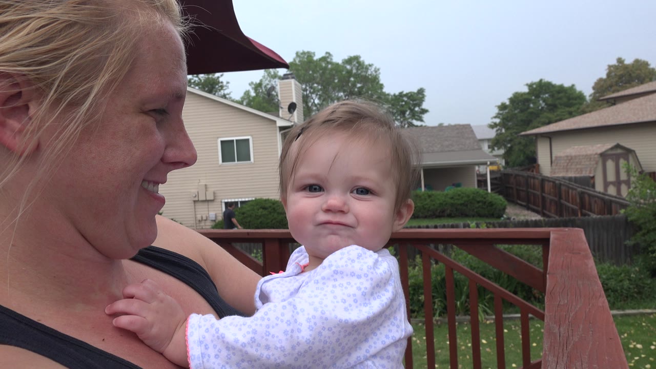 In Westminster, Colorado, the woman turns toward the baby, who sits in her arms near the red deck rail with houses, lawn, and trees beyond.
