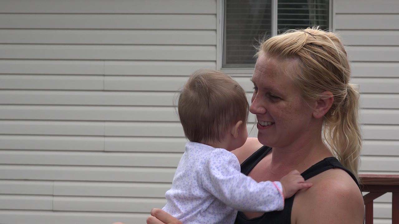 In Westminster, Colorado, a blonde mother smiles at a small child she is holding close on a wooden backyard deck beside beige house siding.