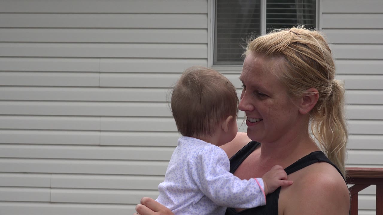 The mother holds the child at chest height as they face each other, with horizontal beige siding and a dark window behind them in Westminster, Colorado.