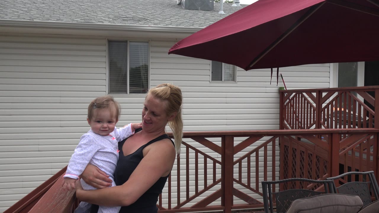 In Westminster, Colorado, the mother and young child remain near the left side of the deck, framed by the red railing, umbrella canopy, and house wall.