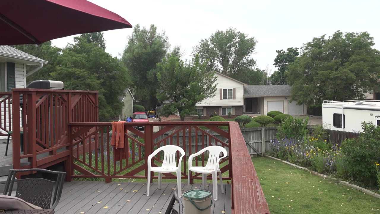 Backyard deck with two white chairs, red railing, lawn, fence, RV, and houses.