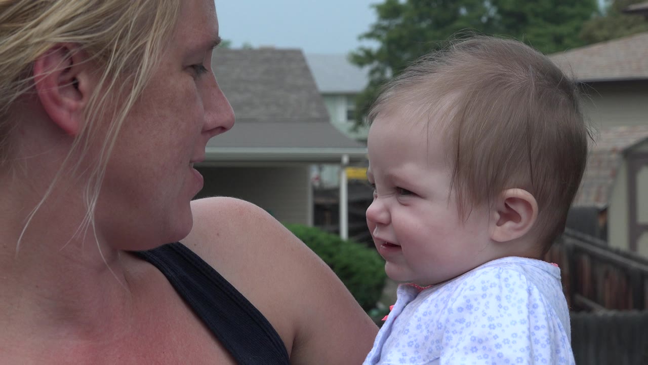 In Westminster, Colorado, a mother in profile holds a toddler outdoors while both look toward each other near a wooden fence and residential buildings.
