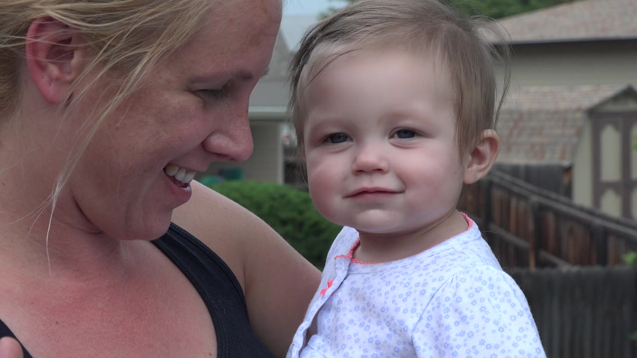 A blonde mother smiles beside a baby girl facing the camera in Westminster, Colorado, with a backyard fence and houses behind them.