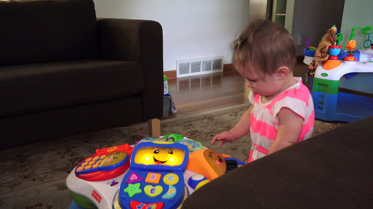 A toddler in a pink striped shirt looks down beside a colorful activity toy in a Westminster, Colorado living room with a dark sofa and floor vent behind.