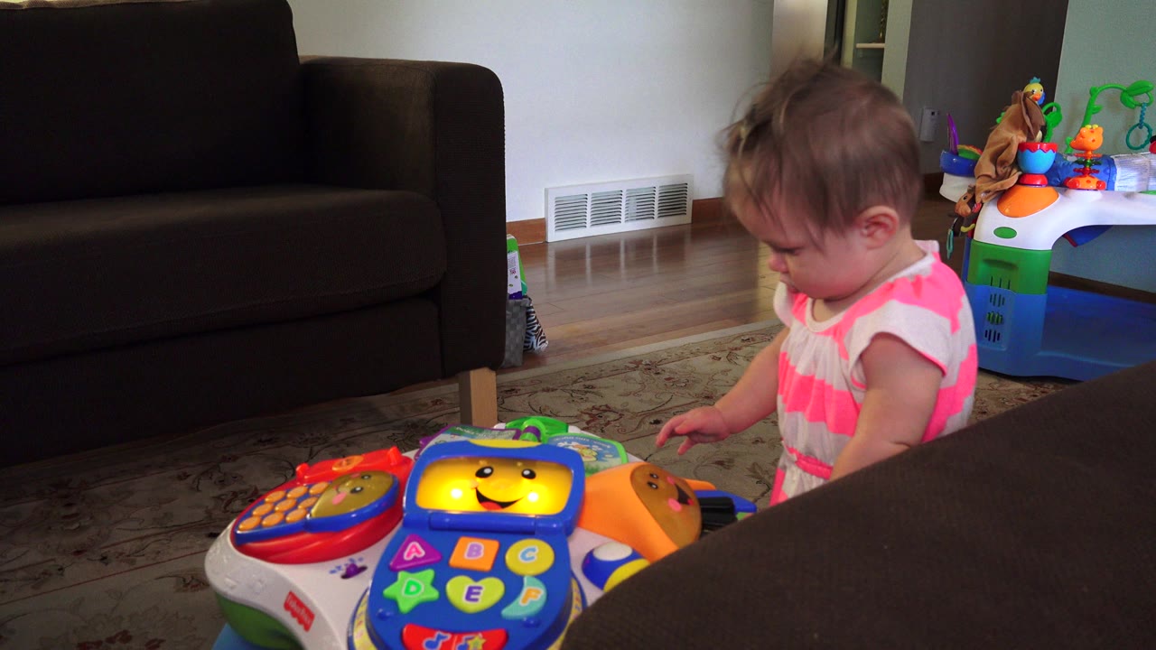 The child keeps looking down at the plastic learning toy, with the dark couch filling the left background and additional toys near the wall in Westminster, Colorado.