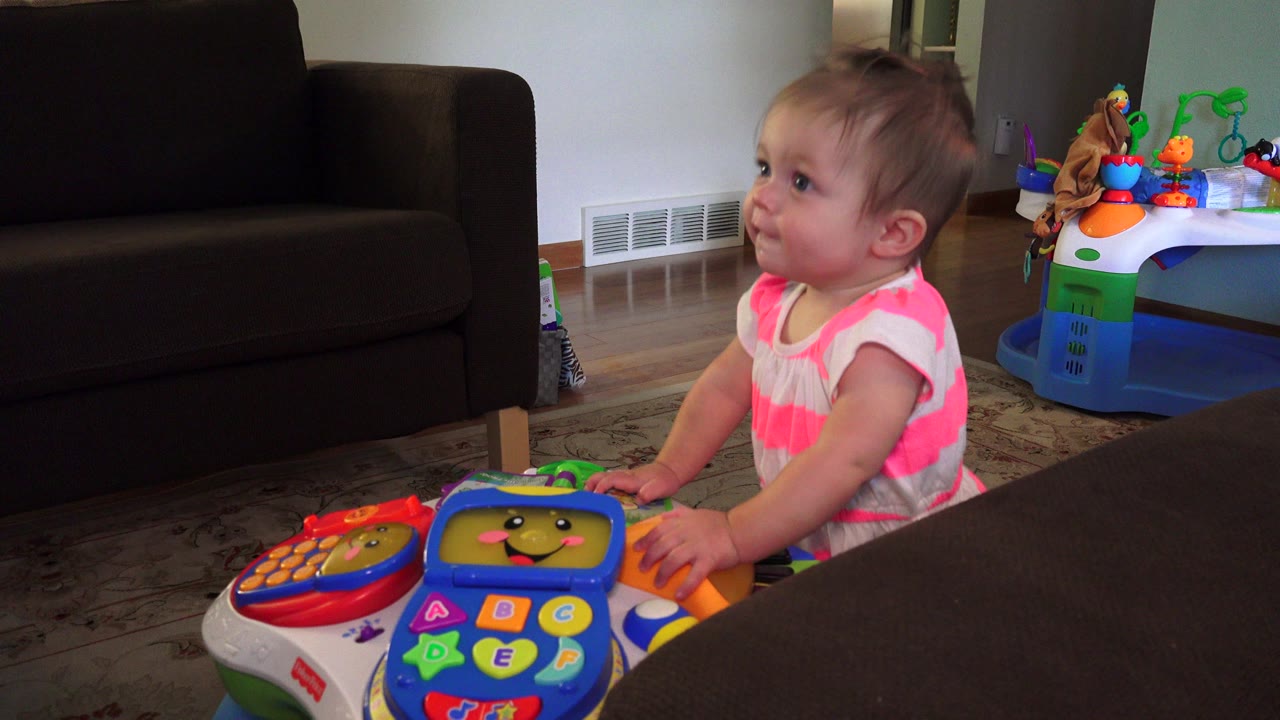 Toddler girl leaning on a colorful electronic playset in a living room.