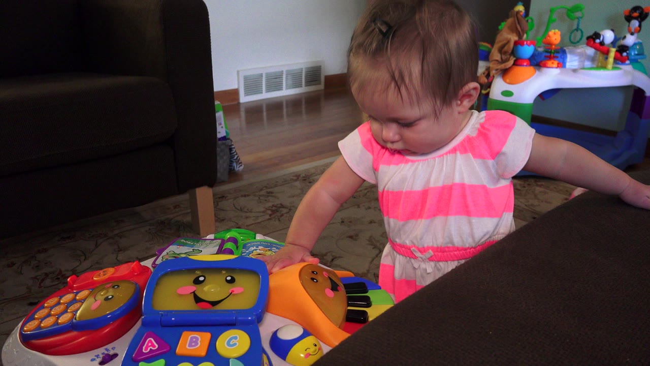 Toddler in a pink striped dress reaches toward colorful toys beside a sofa.