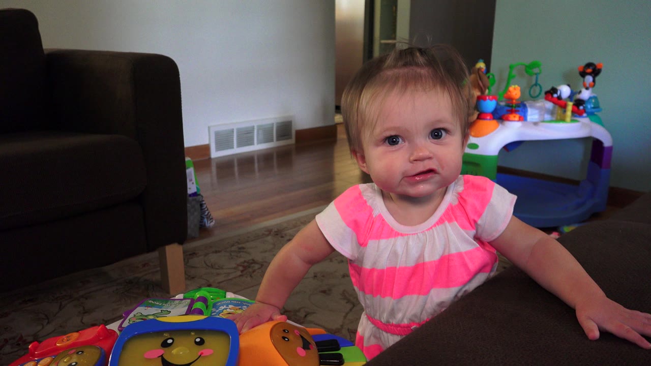 The toddler remains upright beside the dark table in the Westminster home, with colorful play equipment and a dark chair visible in the background.