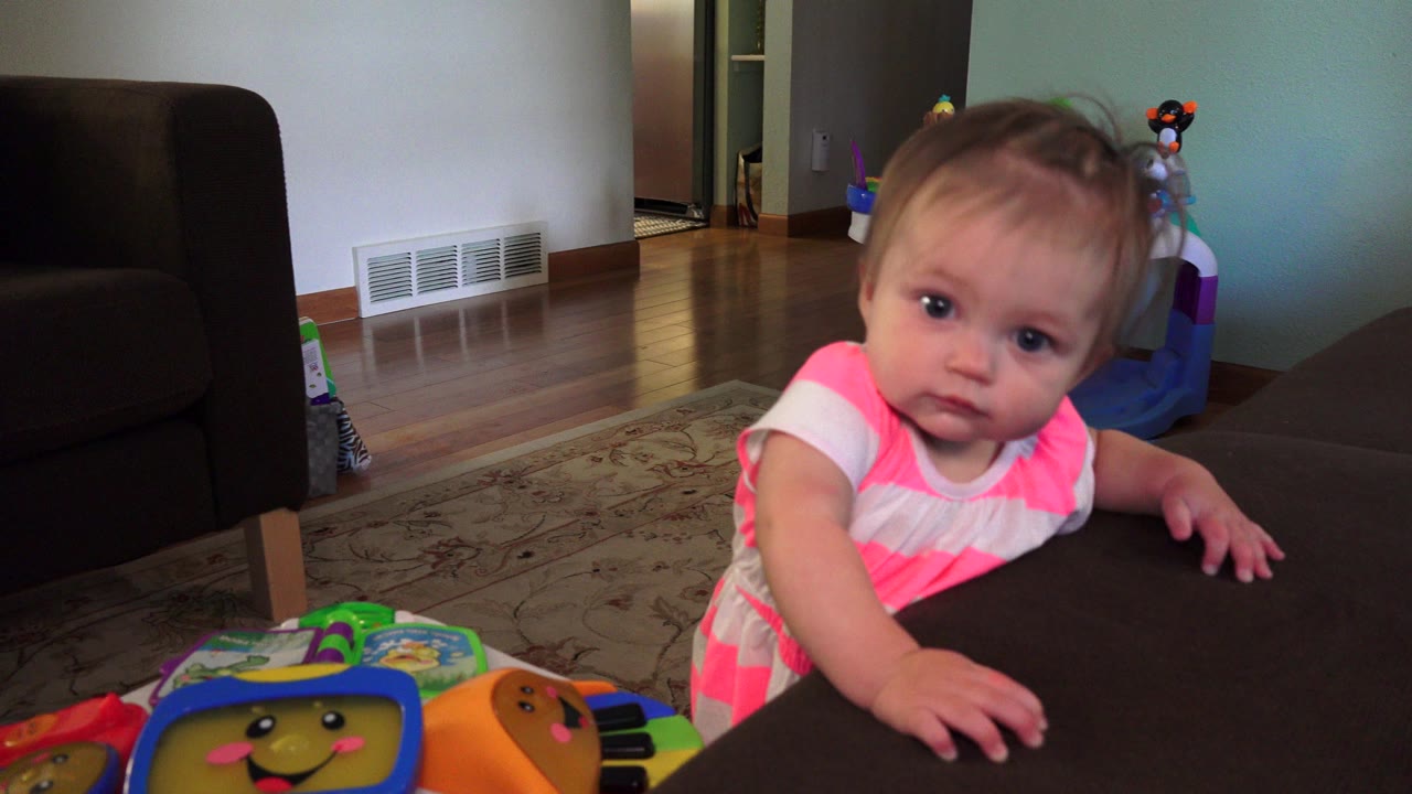 Baby standing beside a dark couch near a colorful toy