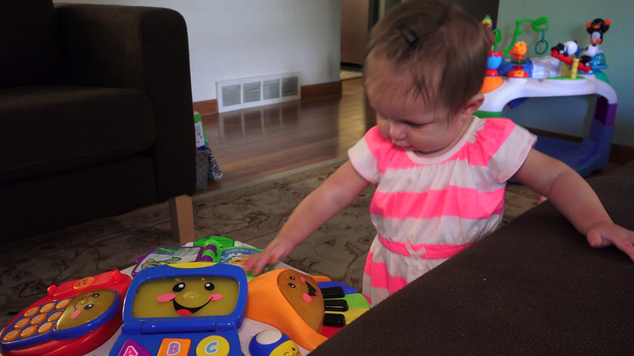 The toddler looks down toward the toy while one hand rests near the couch, with a toy table and wall vent visible behind her in the Westminster, Colorado home.