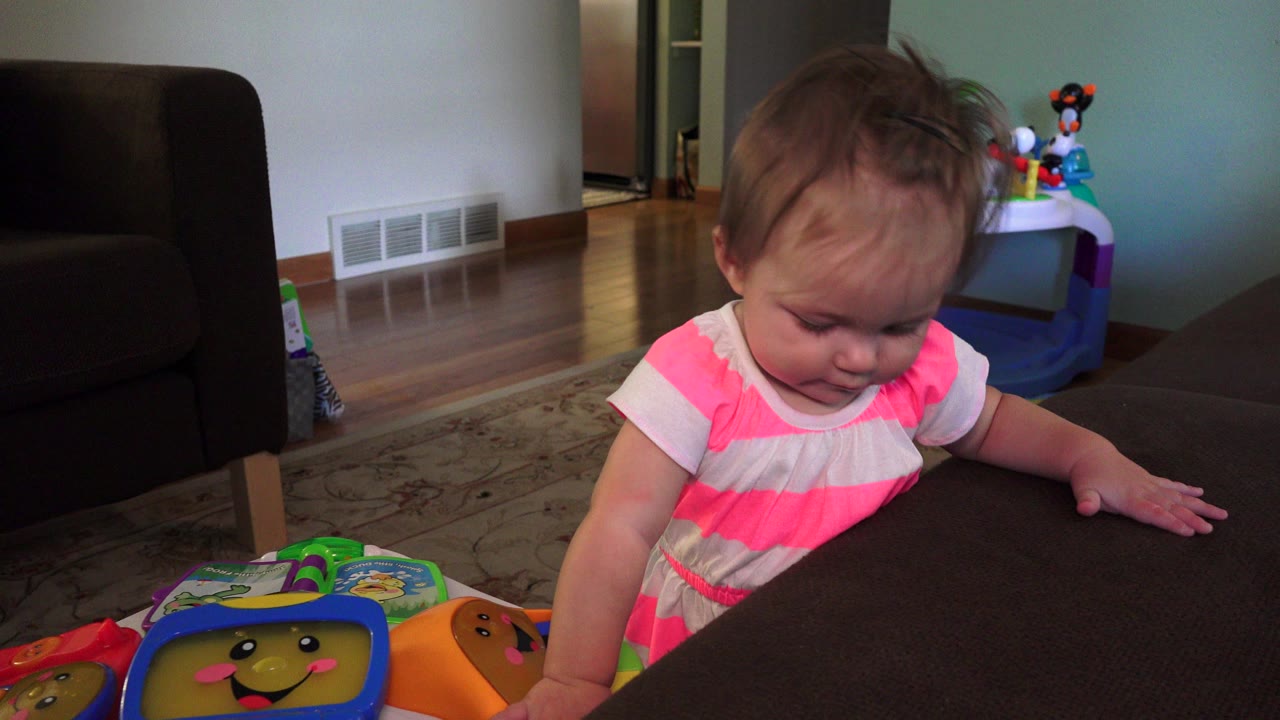 Toddler in a pink and white dress leaning beside a couch and toy.