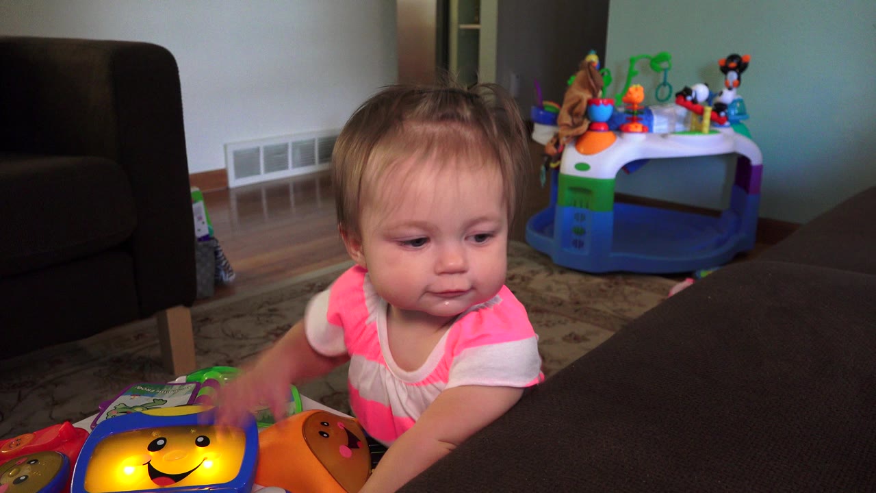 A toddler in a pink and white striped shirt stands near a dark couch and reaches toward a colorful toy in a Westminster, Colorado living room.
