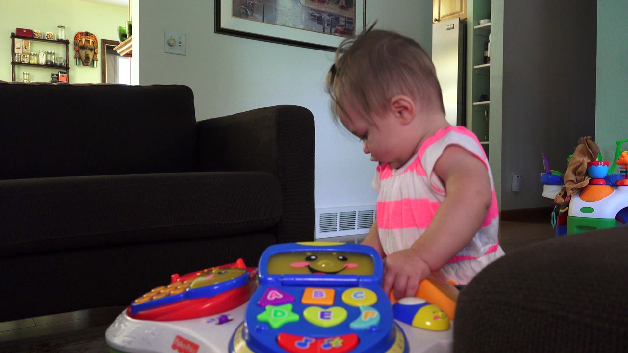 At 0:00, a toddler in a pink-and-white striped top looks down at a colorful toy activity table inside a Westminster, Colorado home.