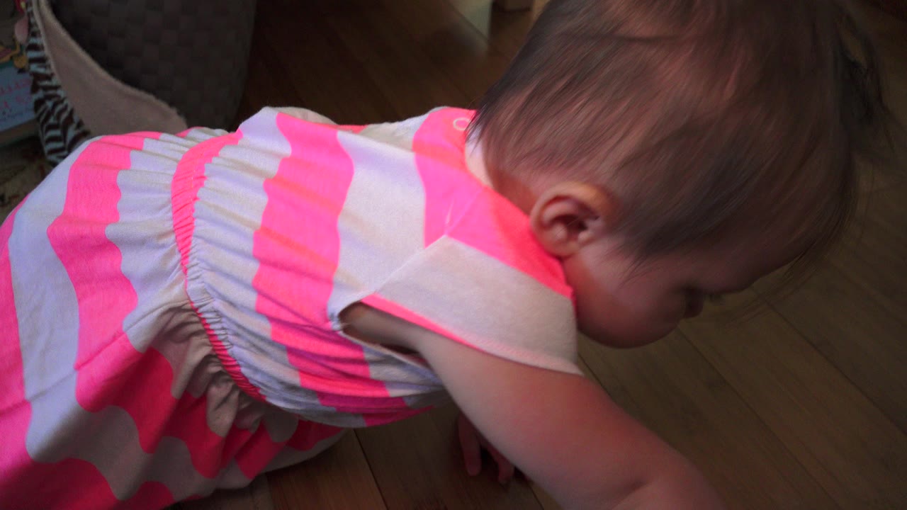 Toddler in a pink and white dress leaning over a hardwood floor.