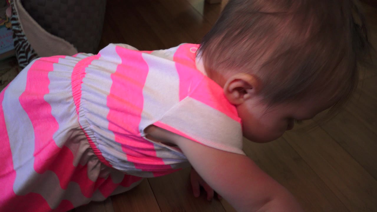 At 0:01, the child remains bent forward in the striped dress, with the hardwood floor and darker room background filling most of the frame.
