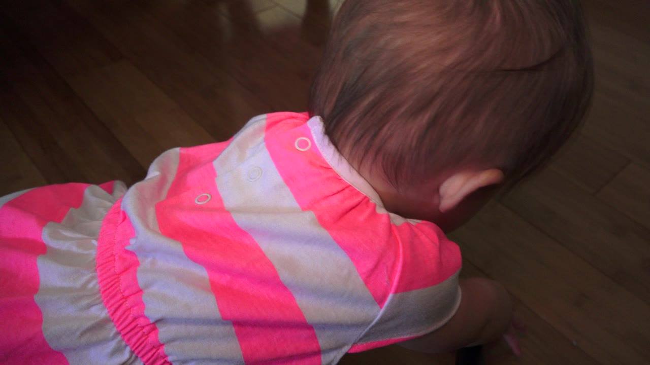 The child remains close to the wooden floor in a Westminster, Colorado home, with the camera looking down at the back of her head and striped outfit.