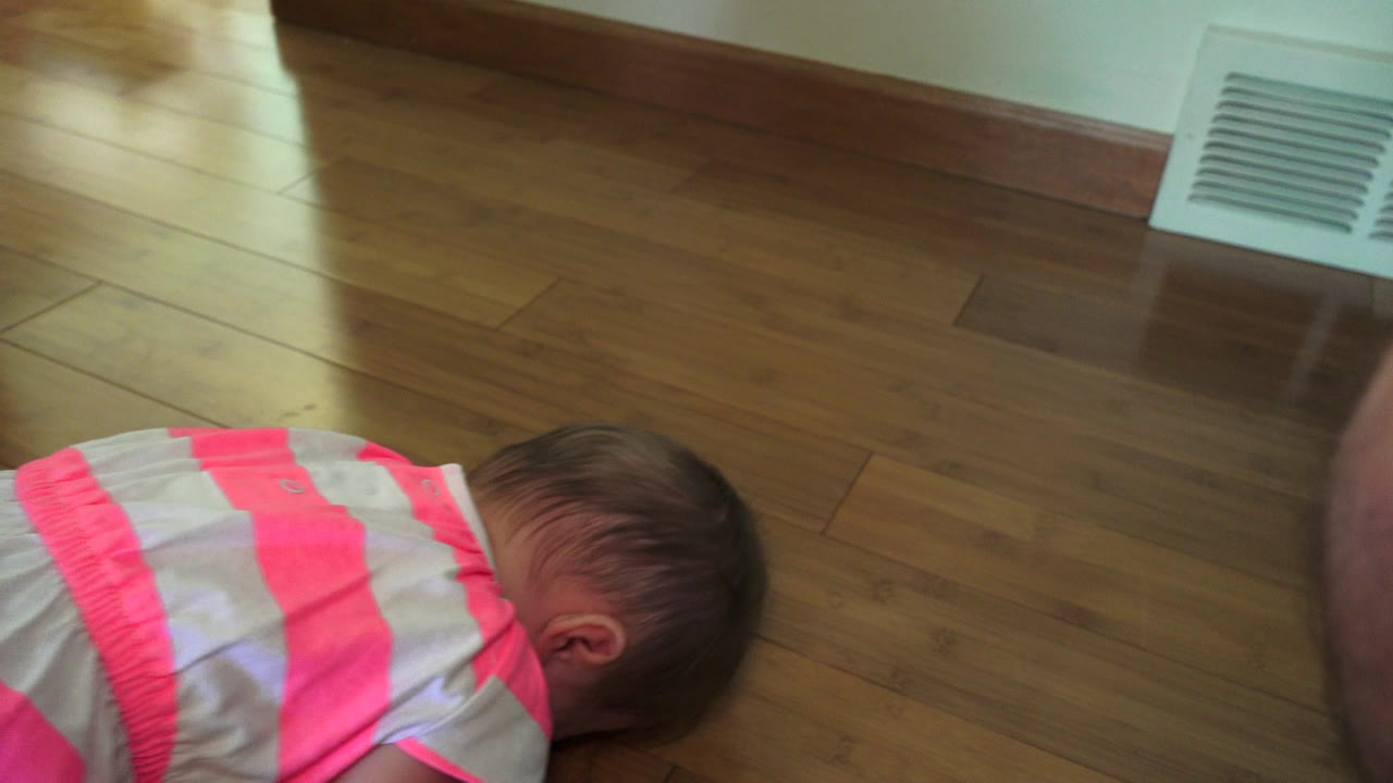 Baby in a striped pink dress low on a wood floor near a wall vent.