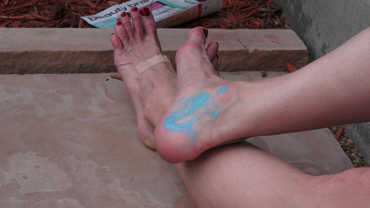 Bare legs and feet rest on concrete steps in Broomfield, Colorado, with red toenails, a toe bandage, blue paint on the heel, mulch, and printed material behind them.