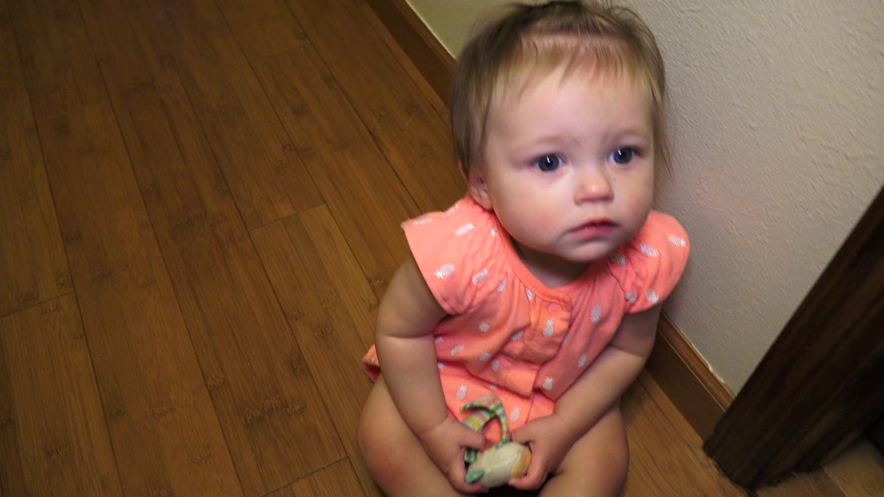 At 0:01, the baby girl keeps her gaze raised toward the camera, with the toy held at her lap and the wood floor filling most of the Broomfield interior frame.