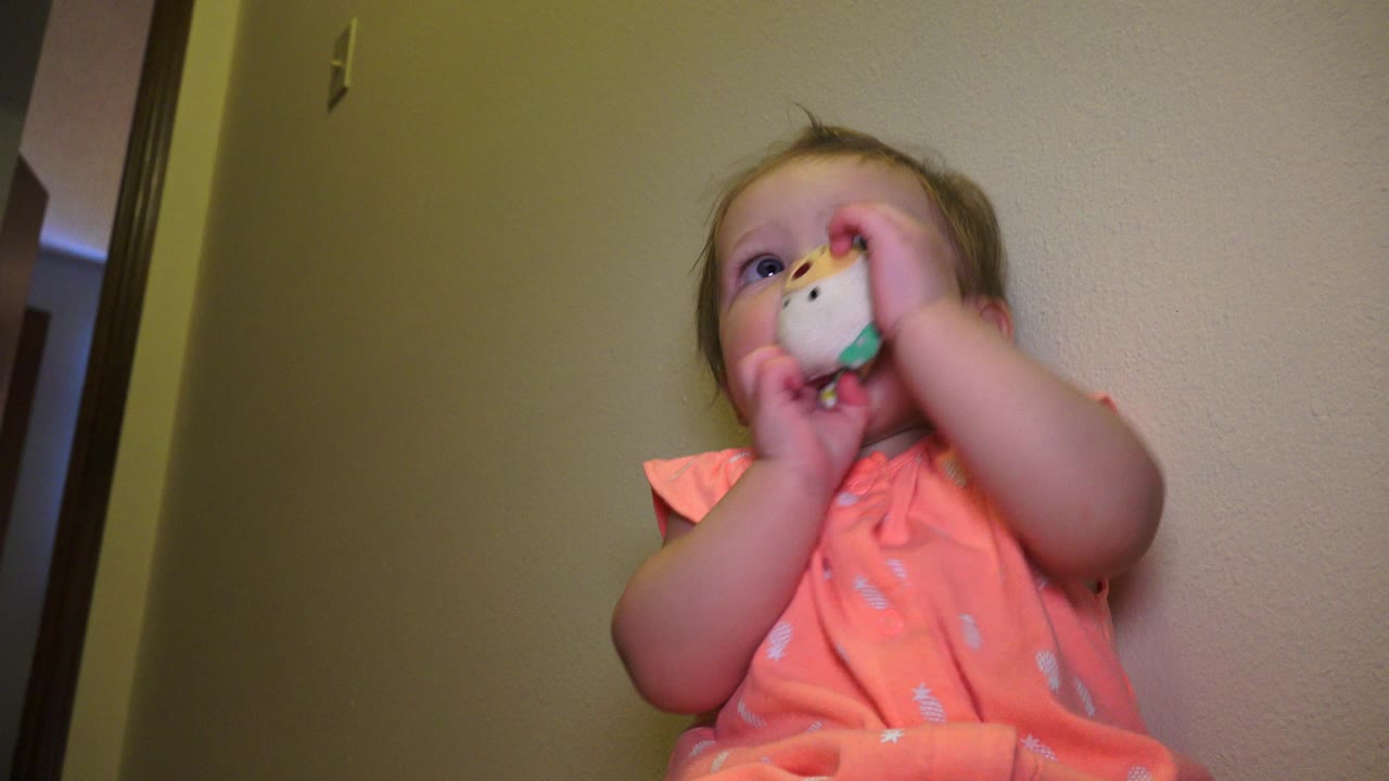 At 0:00 in Broomfield, Colorado, a baby in an orange shirt presses a small white and green object to their mouth while seated against beige interior walls.