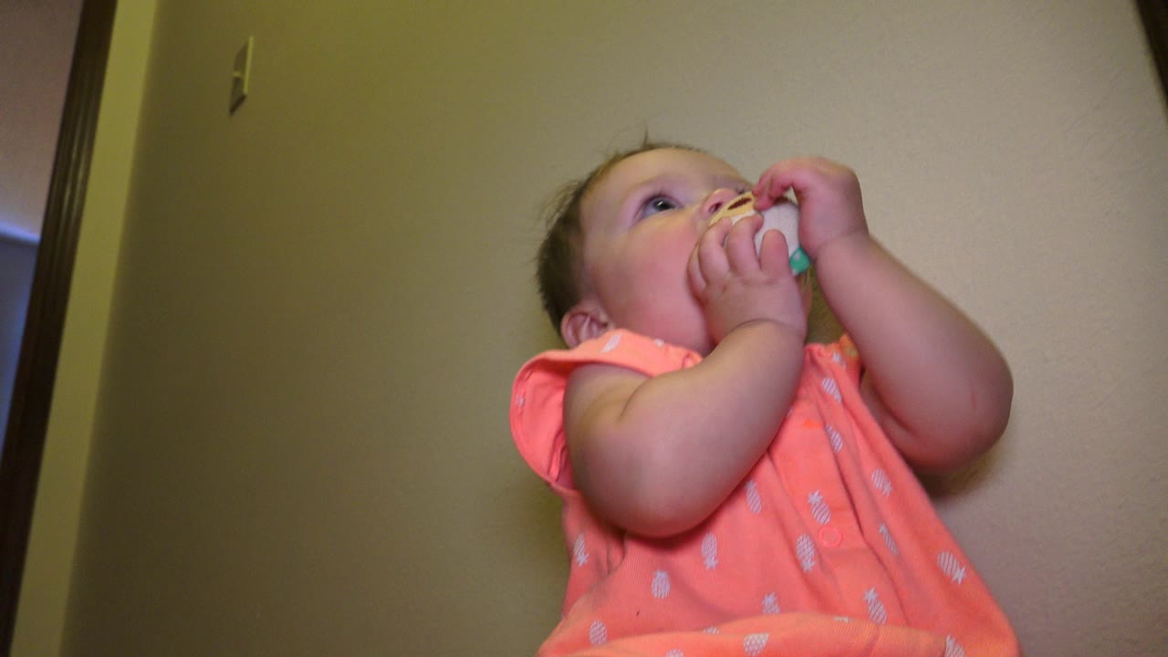 At 0:00 in Broomfield, Colorado, a baby in a coral patterned romper looks upward while holding a small toy to her mouth near beige interior walls.