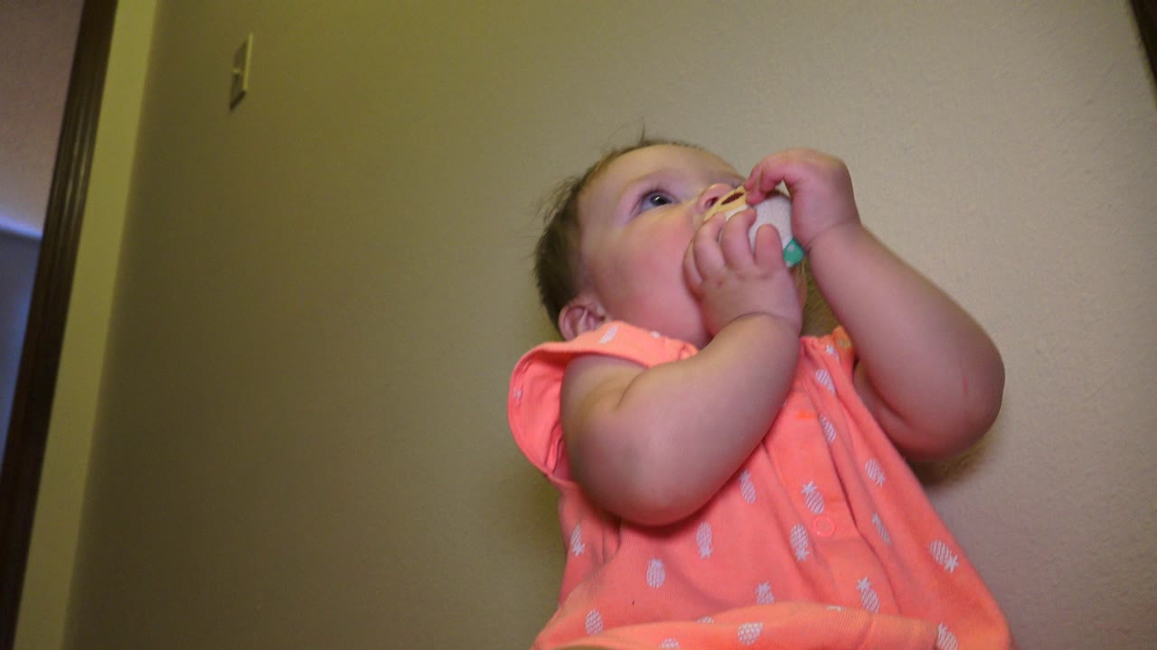 At 0:01 in Broomfield, Colorado, the baby remains close to the camera with both hands raised and the toy partly visible against the beige wall.