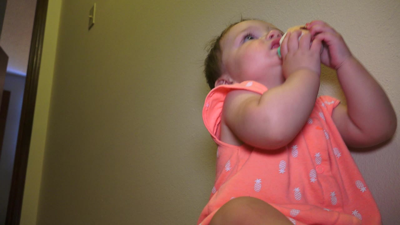 Baby in a coral romper sits at the right side of the frame with a toy at her mouth.