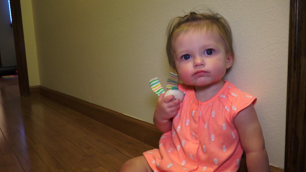 The baby girl remains seated beside the beige wall in Broomfield, Colorado, looking toward the camera with the toy raised in her right hand.