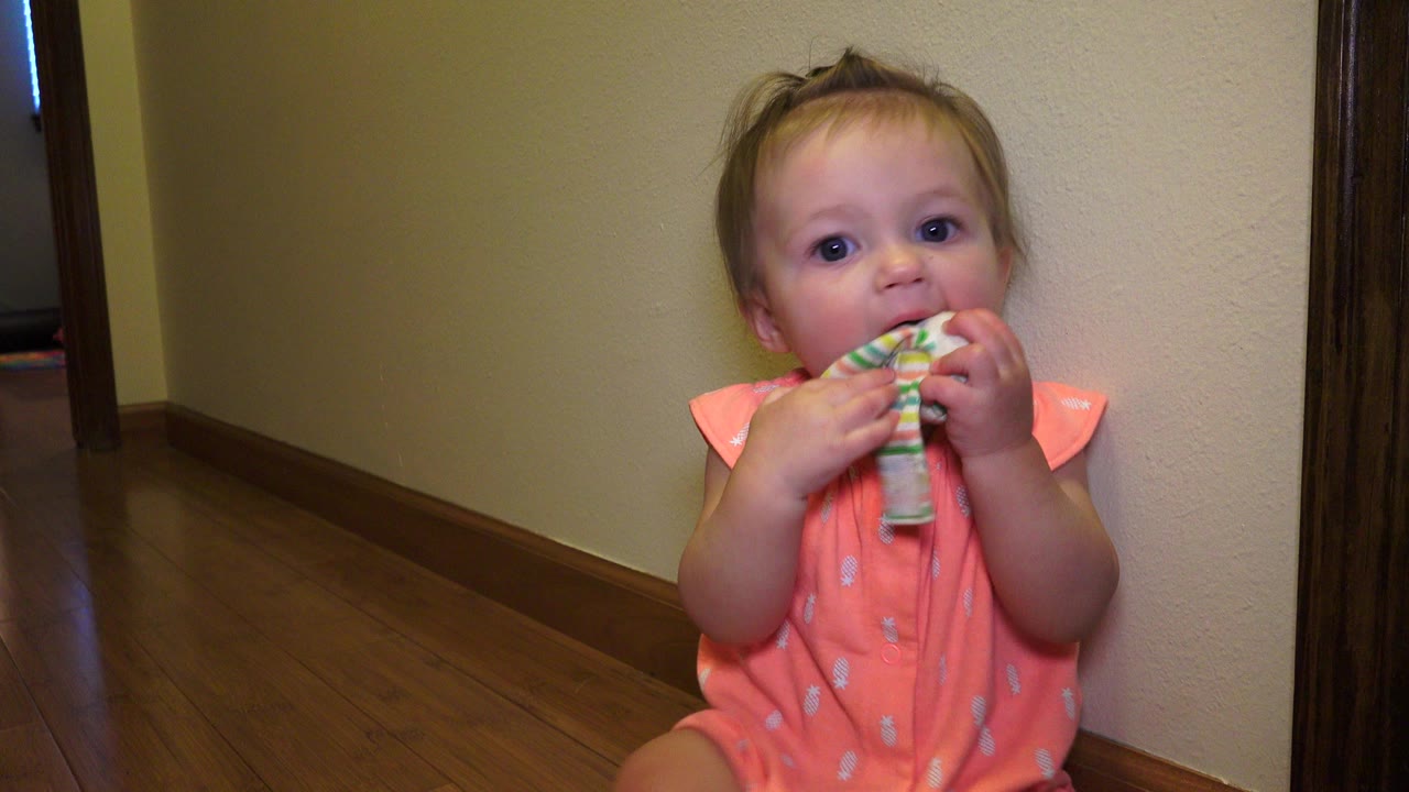 Toddler in an orange dress chewing a rolled pair of socks indoors.