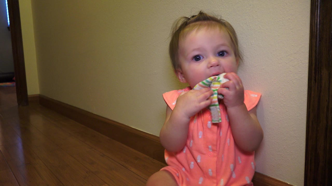 In Broomfield, Colorado, the toddler remains seated close to the beige wall, looking toward the camera with both hands wrapped around the striped socks at her mouth.