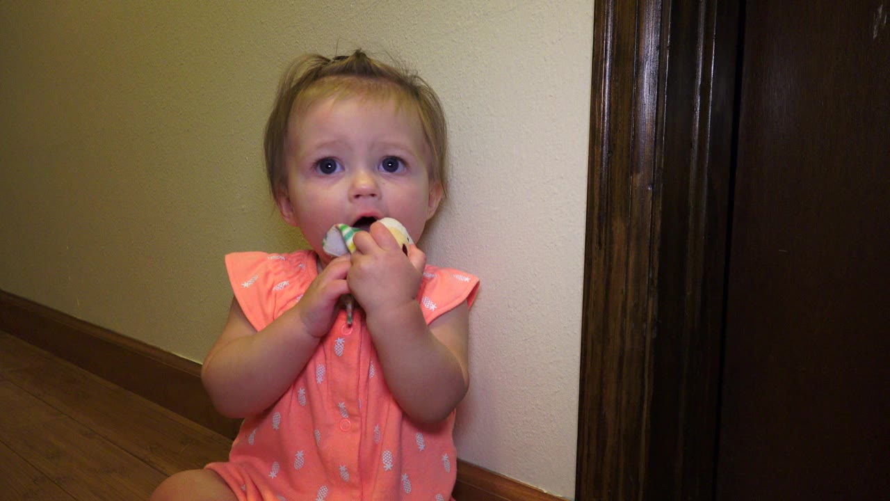 Young child seated by a wall holding a small toy to her mouth.