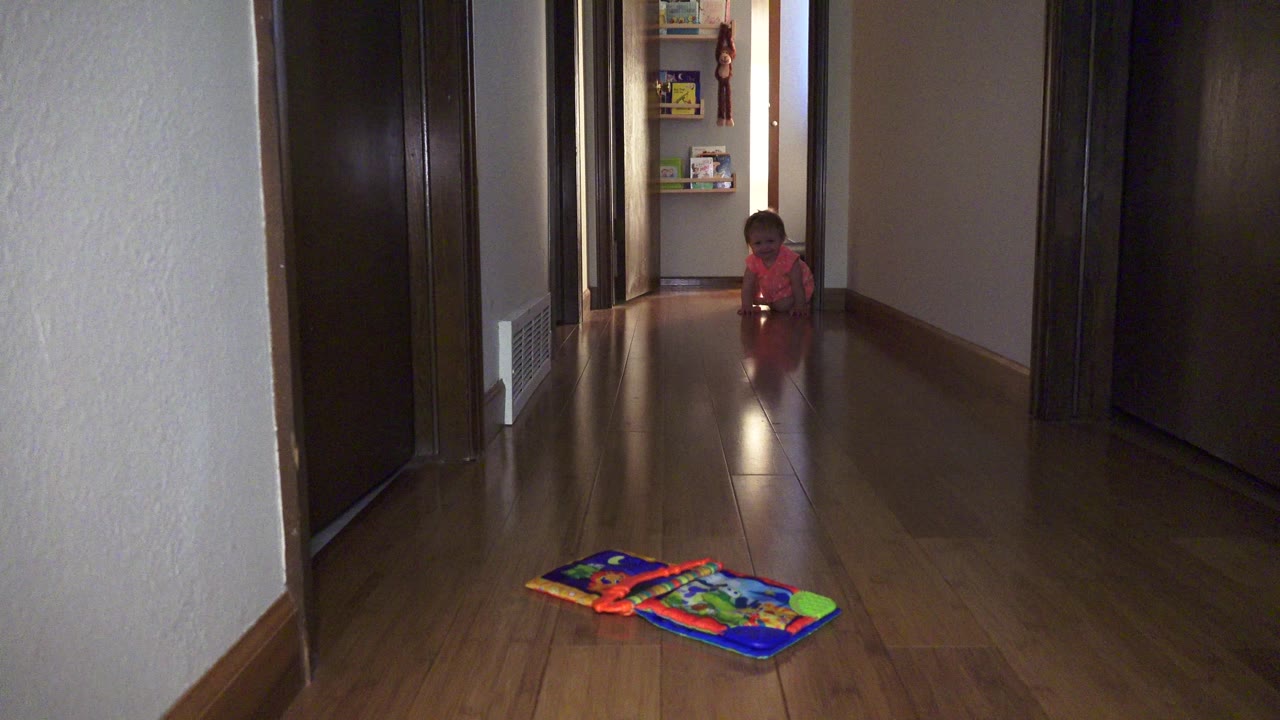 At 0:01 in Broomfield, Colorado, the toddler stays near the far doorway while the bright toy mat sits on the reflective hardwood floor in the foreground.