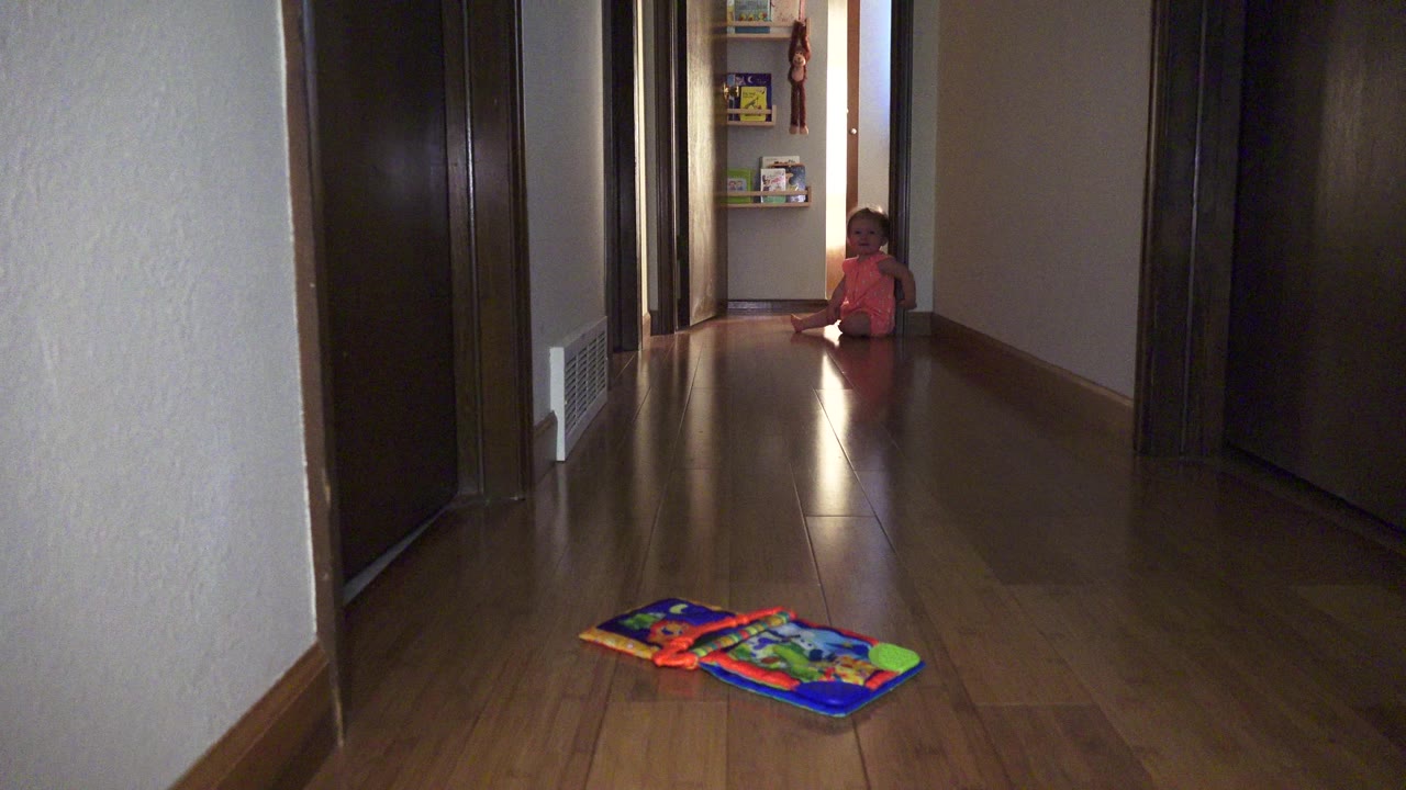 The child remains seated near the end of the Broomfield hallway, with warm light reflecting along the wood floor between the foreground toy mat and her position.