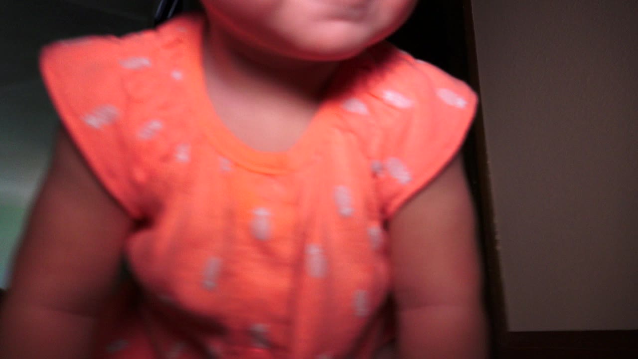 A toddler fills the foreground in a Broomfield, Colorado interior, with an orange shirt, partial face, dark trim, and a lit room behind.