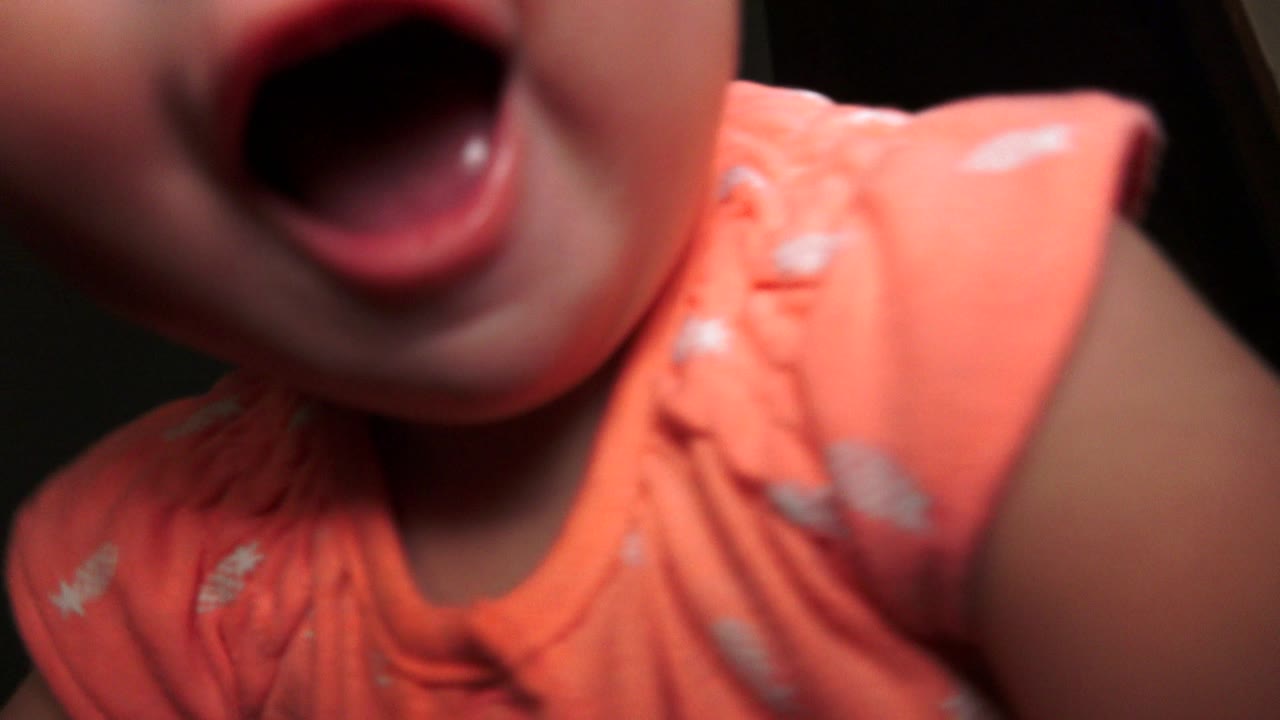 At 0:01 in Broomfield, Colorado, the baby remains close to the lens with the mouth open, shoulder and orange shirt prominent against a dark indoor background.